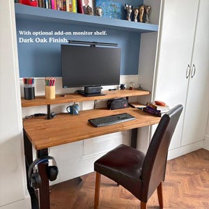 Solid wood desk with a monitor shelf, medium to dark wood finish and black steel legs. Placed in a narrow alcove with a few book shelves above it.