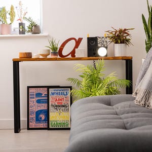 Wooden console table with black square metal legs, styled as a radiator shelf in a modern hallway with plants, books, and decorative objects on top.