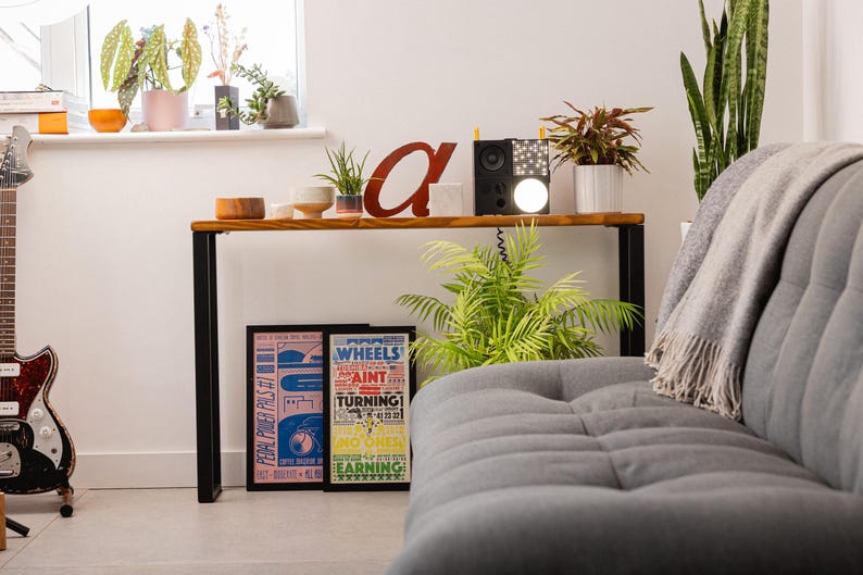Wooden console table with black square metal legs, styled as a radiator shelf in a modern hallway with plants, books, and decorative objects on top.