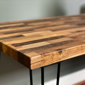 May include: A wooden table top with a unique parquet design, featuring various shades of brown wood. The table is supported by four black hairpin legs.