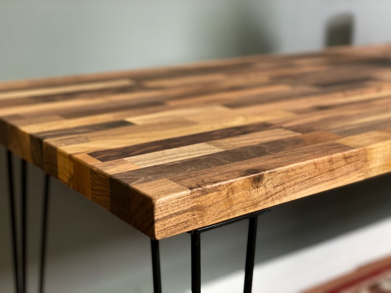 May include: A wooden table top with a unique parquet design, featuring various shades of brown wood. The table is supported by four black hairpin legs.