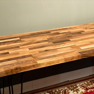 May include: A wooden table top with a natural wood finish. The table top is made of multiple pieces of wood that are arranged in a parquet pattern. The table top is supported by black hairpin legs.