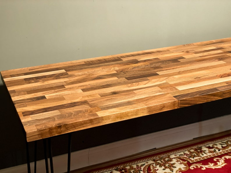 May include: A wooden table top with a natural wood finish. The table top is made of multiple pieces of wood that are arranged in a parquet pattern. The table top is supported by black hairpin legs.