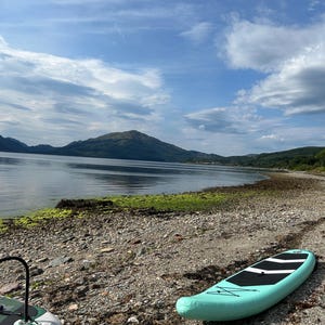 May include: A light mint green stand-up paddleboard with a black traction pad and black bungee cord rigging rests on a rocky shoreline. A second partially visible paddleboard with a green patterned surface sits nearby on the pebble beach.