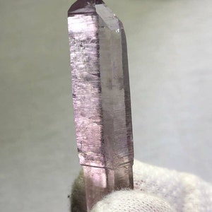 Puede incluir: Cristal translúcido de color púrpura claro con una punta afilada. El cristal tiene forma rectangular con estrías visibles a lo largo. Lo sostiene una mano enguantada, posiblemente un espécimen o una piedra preciosa.