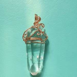 Puede incluir: Colgante de cristal de cuarzo transparente envuelto en alambre de cobre. El cristal está facetado y tiene una punta. El alambre de cobre está intrincadamente moldeado alrededor de la parte superior del cristal, formando una argolla decorativa para un collar.