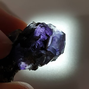Puede incluir: Primer plano de un grupo de cristales de fluorita. El cristal presenta un tono púrpura intenso con formas geométricas translúcidas. La base del grupo es de un color oscuro y terroso, que contrasta con los vibrantes cristales púrpuras.