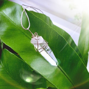 Puede incluir: Un colgante de cristal de cuarzo transparente, envuelto en alambre plateado, suspendido de un cordón blanco. El cristal se muestra sobre un fondo de hojas verdes grandes y vibrantes. Un accesorio natural y terroso.