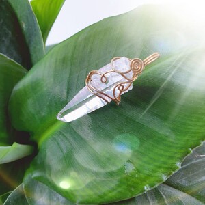Peut inclure: Un pendentif en cristal de quartz clair, enveloppé dans du fil de cuivre, posé sur une grande feuille verte. Le fil est façonné en spirales décoratives et en boucle pour suspendre. Le cristal est pointu et facetté.