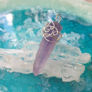 Peut inclure: Pendentif en cristal d'améthyste violet avec un fil d'argent. Le fil est conçu de manière complexe autour du cristal, formant un motif décoratif. Le pendentif est présenté sur un lit de cristaux clairs sur fond turquoise. Environ 7 cm de long.