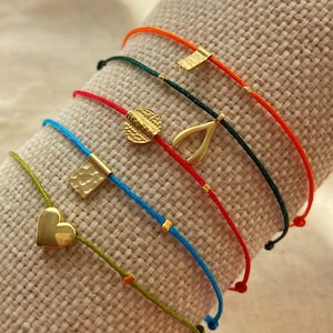May include: A set of colourful string bracelets with gold charms. The charms include a heart, a horseshoe, a square, a circle and a rectangle.