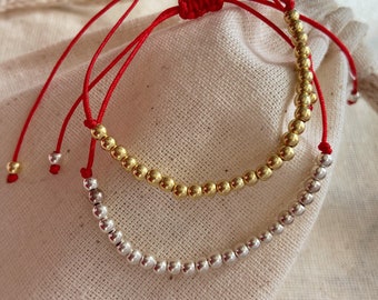May include: Two bracelets with red string and beads. One bracelet has gold beads and the other has silver beads.