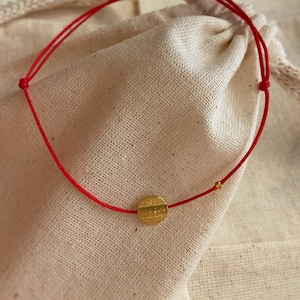 Può includere: Un braccialetto con filo rosso con un piccolo ciondolo a disco dorato strutturato e una minuscola perla dorata. Il braccialetto è presentato su un sacchetto beige con coulisse, creando un'estetica minimalista.