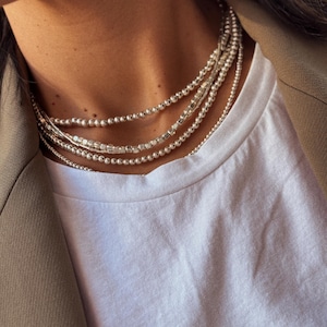 May include: A close-up of a person wearing a layered silver beaded necklace. The necklace is made of small, round beads and is worn on a white t-shirt.