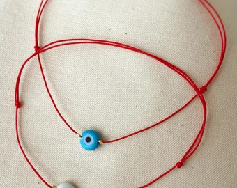 May include: Two red string bracelets with evil eye charms. One charm is blue with a black pupil and the other is white with a black pupil.