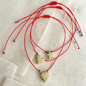 Puede incluir: Tres pulseras de cuerda roja con dijes de oro. Los dijes son un corazón, una estrella y un trébol de cuatro hojas. Las pulseras están atadas con cuentas azules.