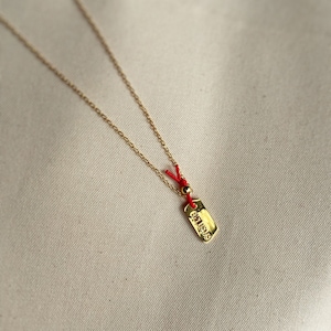 May include: A gold necklace with a small gold tag pendant that says "BELIEVE" in all caps. The necklace is on a white background and has a red string attached to the tag.
