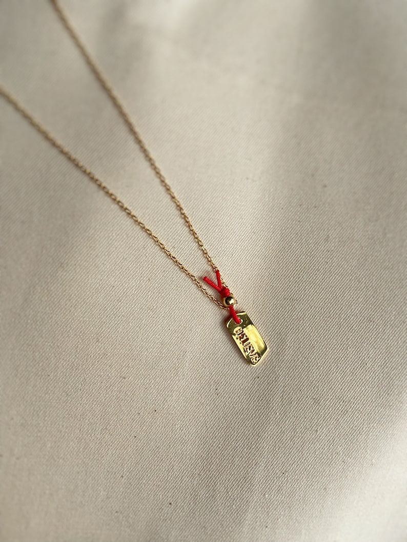 May include: A gold necklace with a small gold tag pendant that says "BELIEVE" in all caps. The necklace is on a white background and has a red string attached to the tag.