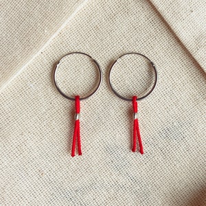 May include: A pair of silver hoop earrings with red tassel charms hanging from them. The earrings are on a beige fabric background.