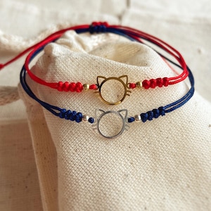 May include: Two cat-shaped charm bracelets. One bracelet has a red cord with a gold cat charm and gold beads. The other bracelet has a blue cord with a silver cat charm and silver beads. Both bracelets are on a white cloth.