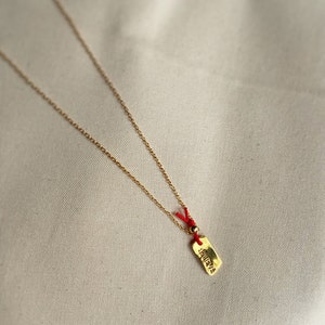 May include: A gold chain necklace with a small gold tag pendant. The tag has the word "believe" written on it in a stylized font. The chain is attached to the tag with a red string and a small gold bead.