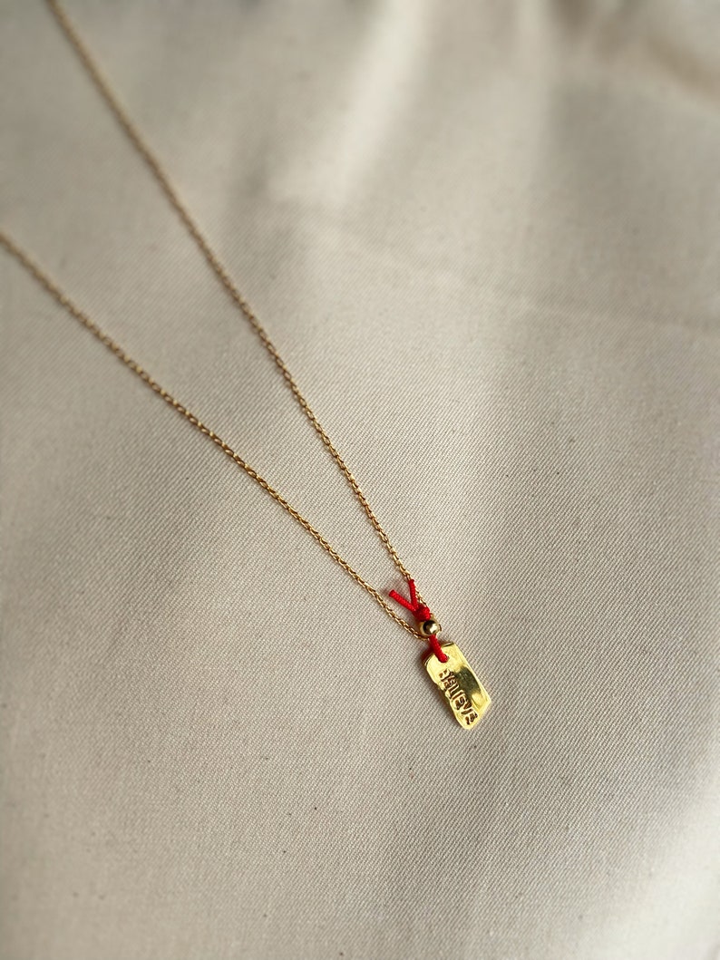 May include: A gold chain necklace with a small gold tag pendant. The tag has the word "believe" written on it in a stylized font. The chain is attached to the tag with a red string and a small gold bead.
