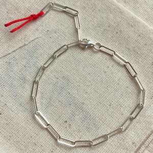 Pode incluir: Um bracelete de corrente de prata com fecho de encaixe e um cord&atilde;o vermelho preso &agrave; extremidade. O bracelete &eacute; feito de pequenos elos retangulares.