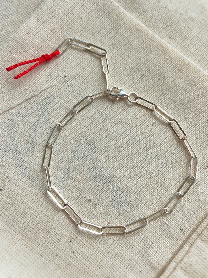 Pode incluir: Um bracelete de corrente de prata com fecho de encaixe e um cord&atilde;o vermelho preso &agrave; extremidade. O bracelete &eacute; feito de pequenos elos retangulares.