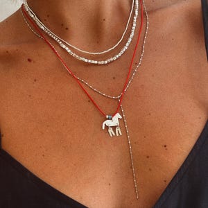 May include: A layered necklace set consisting of two silver-toned chains with small metallic beads and a red cord necklace featuring a silver-toned horse pendant with two dangling rectangular charms and a small blue bead accent at the top.