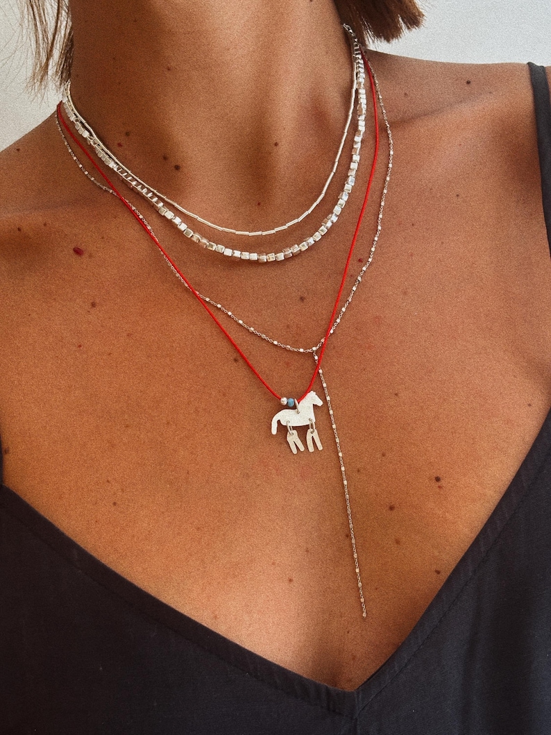 May include: A layered necklace set consisting of two silver-toned chains with small metallic beads and a red cord necklace featuring a silver-toned horse pendant with two dangling rectangular charms and a small blue bead accent at the top.