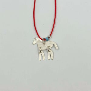 May include: A silver-toned horse pendant suspended from a thin red cord. The horse figure features a textured surface and is attached to the cord by a silver jump ring. A small light blue bead and a tiny silver sphere are threaded onto the red cord.