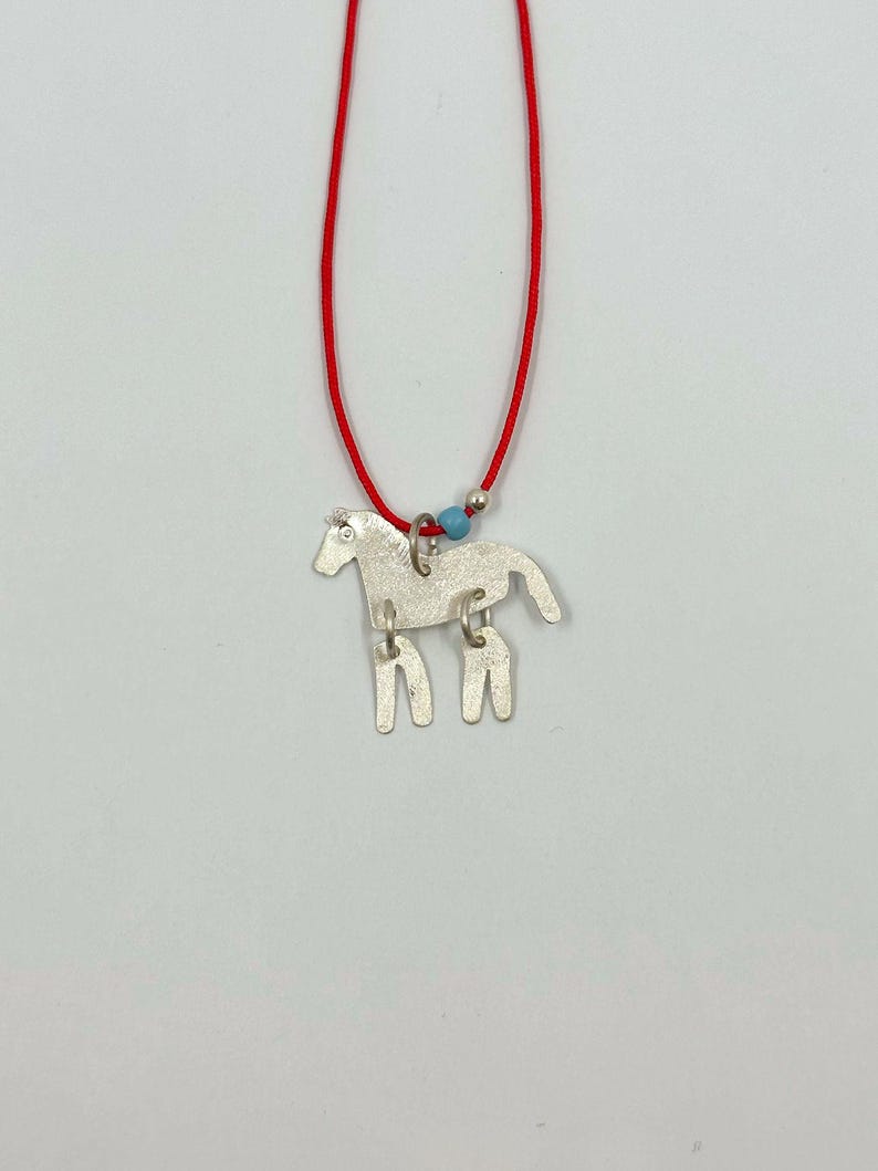 May include: A silver-toned horse pendant suspended from a thin red cord. The horse figure features a textured surface and is attached to the cord by a silver jump ring. A small light blue bead and a tiny silver sphere are threaded onto the red cord.