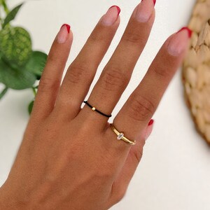 May include: A hand wearing two rings: a thin black cord ring with a gold bead and a gold band ring with a small, rectangular clear stone. The nails are painted with a French manicure style with red tips.