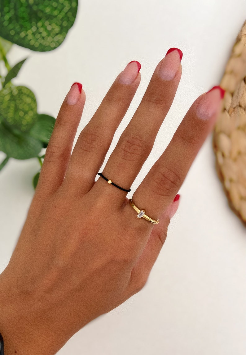 May include: A hand wearing two rings: a thin black cord ring with a gold bead and a gold band ring with a small, rectangular clear stone. The nails are painted with a French manicure style with red tips.