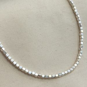 Peut inclure: Un collier argenté composé de petites perles carrées. Les perles sont enfilées pour former une chaîne délicate. Le collier est présenté sur un fond neutre, soulignant son élégance simple. Un bijou minimaliste.