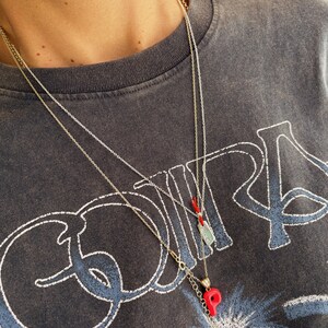 May include: Two gold chain necklaces with pendants. The top necklace has a small rectangular pendant with a red string. The bottom necklace has a red letter "P" pendant.