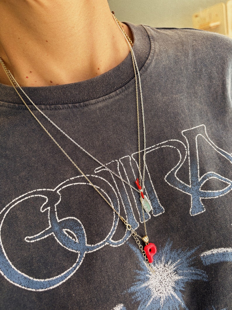May include: Two gold chain necklaces with pendants. The top necklace has a small rectangular pendant with a red string. The bottom necklace has a red letter "P" pendant.