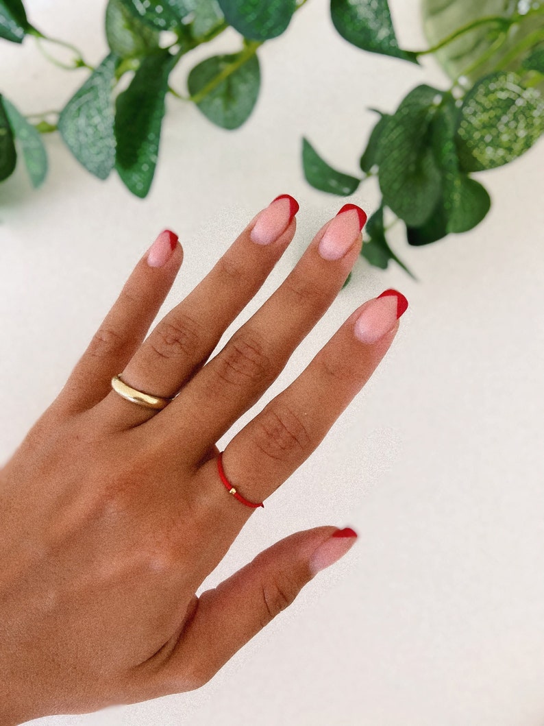 May include: Close-up of a hand with French tip nails featuring a red border. A gold ring and a red string ring with a small gold bead are also visible. Green leaves are in the background.