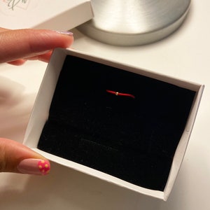 May include: A red string bracelet with a small gold bead, presented in a white box with a black velvet interior. The box has the brand name "Pheia" printed on the lid. The bracelet is a delicate piece of jewelry.