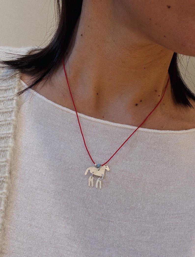 May include: A silver-toned horse pendant necklace featuring a thin red cord. The horse charm has a textured surface and is accented with two small dangling silver-toned shapes attached to its underside, along with a single small blue bead.