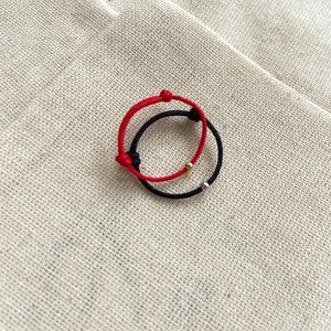 May include: Two adjustable bracelets. One is red, the other is black. Each bracelet has a sliding knot closure and a small metallic bead. The bracelets are displayed on a textured, beige fabric.