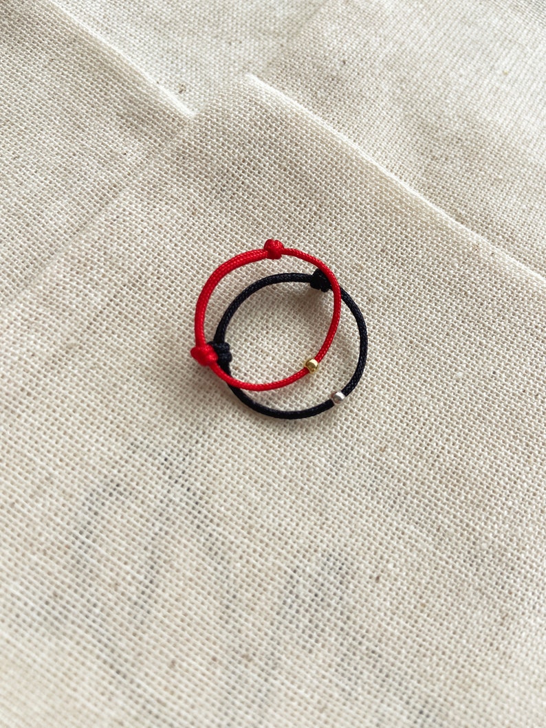 May include: Two adjustable bracelets. One is red, the other is black. Each bracelet has a sliding knot closure and a small metallic bead. The bracelets are displayed on a textured, beige fabric.