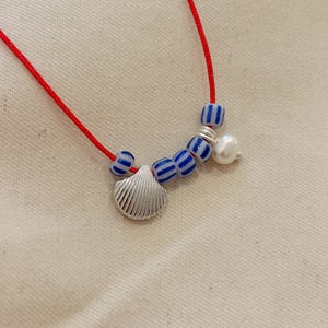 May include: A necklace featuring a thin red cord strung with four small blue and white striped beads, a silver-toned metal seashell charm, and a single white freshwater pearl accent. The components are arranged along the cord in a linear fashion.