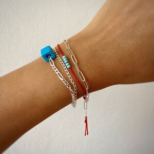 Pode incluir: Um conjunto de tr&ecirc;s pulseiras em um pulso. As pulseiras s&atilde;o uma corrente de prata, uma pulseira de cord&atilde;o vermelho com um fecho de prata e uma pulseira de contas turquesa.