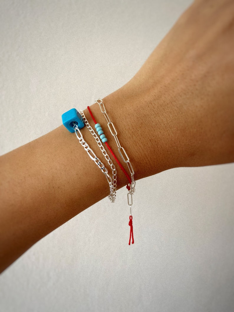Pode incluir: Um conjunto de tr&ecirc;s pulseiras em um pulso. As pulseiras s&atilde;o uma corrente de prata, uma pulseira de cord&atilde;o vermelho com um fecho de prata e uma pulseira de contas turquesa.