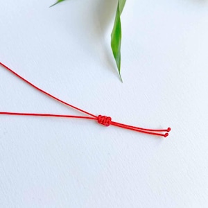 May include: A thin, vibrant red cord featuring a central sliding knot mechanism. The cord is constructed from smooth, durable thread, with two loose ends extending from the adjustable knot, designed for a bracelet or necklace closure.