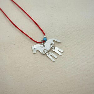 May include: A silver-toned metal horse pendant necklace featuring a red cord. The horse charm has a textured surface and is accented with a small, round, light blue bead. The pendant is suspended from a thin, vibrant red cord necklace.