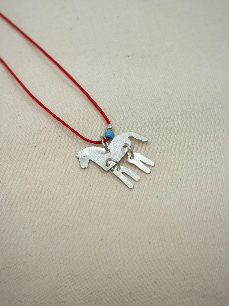 May include: A silver-toned metal horse pendant necklace featuring a red cord. The horse charm has a textured surface and is accented with a small, round, light blue bead. The pendant is suspended from a thin, vibrant red cord necklace.