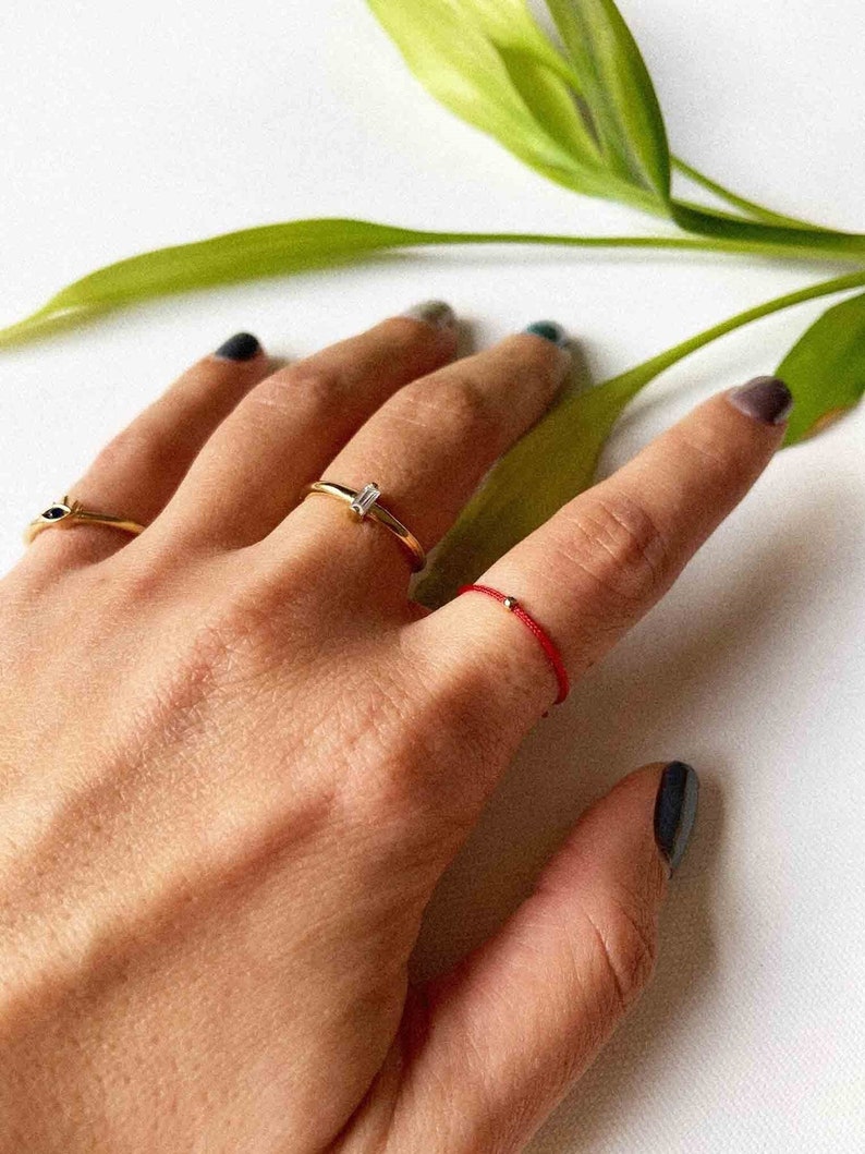 May include: A hand displays three rings: a gold band with a small black stone, a gold band with a rectangular clear stone, and a red string ring with a small gold bead. Green leaves are in the background.
