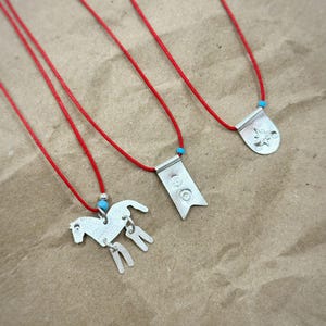 May include: Three necklaces featuring silver-toned metal pendants on thin red cords. One pendant is shaped like a horse with dangling legs, one is a rectangular flag with two circular engravings, and one is a rounded shield with a star design.